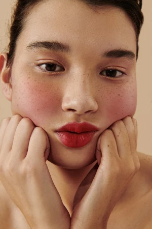 Close-up portrait of a woman with red lipstick and freckles holding her face in her hands against a neutral beige background